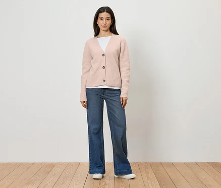 Frau mit rosa Strickjacke, weißem T-Shirt, blauen Jeans und weißen Schuhen steht vor einer weißen Wand.