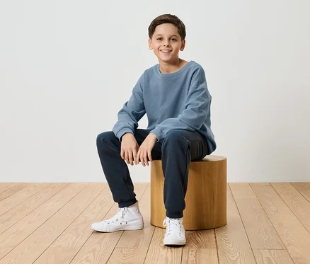 Ein lächelnder Junge sitzt auf einem Holzhocker, gekleidet in einen blauen Pullover, marineblaue Jogginghose und weiße Turnschuhe.