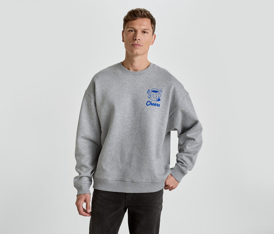 Mann trägt ein graues Sweatshirt mit dem blauen Aufdruck Cheers und einer Tasse darauf.