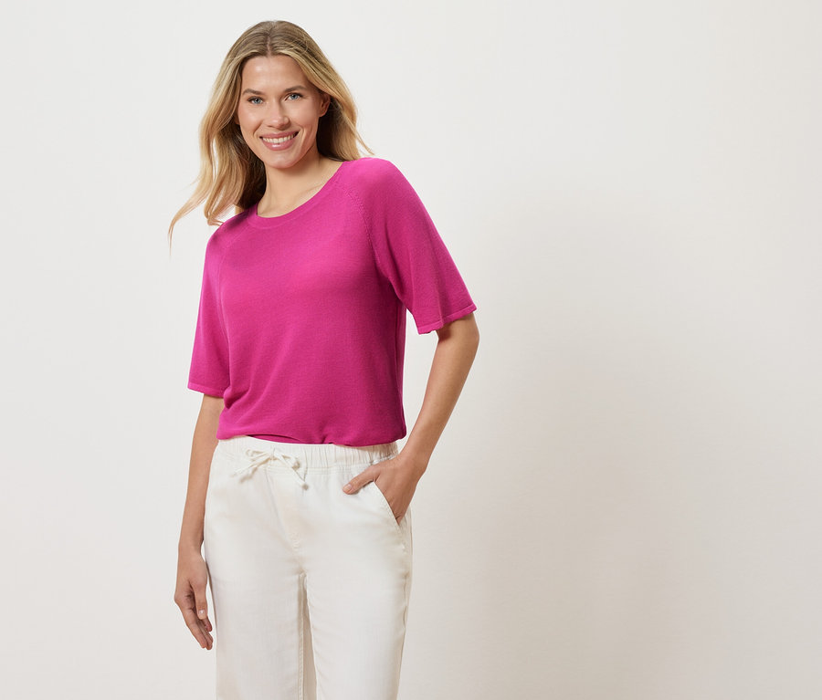 Frau mit blonden, gewellten Haaren, die ein pinkes T-Shirt und weiße Hosen trägt und eine Hand in der Hosentasche hat.