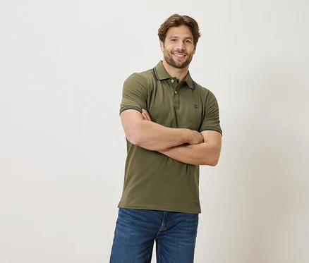 Ein Mann mit verschränkten Armen trägt ein grünes Poloshirt und Jeans vor einem weißen Hintergrund.