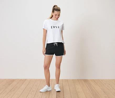 Frau steht in einem weißen T-Shirt mit der Aufschrift LOVE und schwarzen Shorts mit weißen Akzenten.