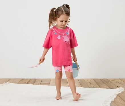 Ein kleines Mädchen mit Zöpfen steht barfuß auf einem weißen Teppich. Sie trägt ein pinkes T-Shirt mit Muscheldekor und rosa Shorts mit Sternen. In ihren Händen hält sie eine Schaufel und einen blauen Eimer.