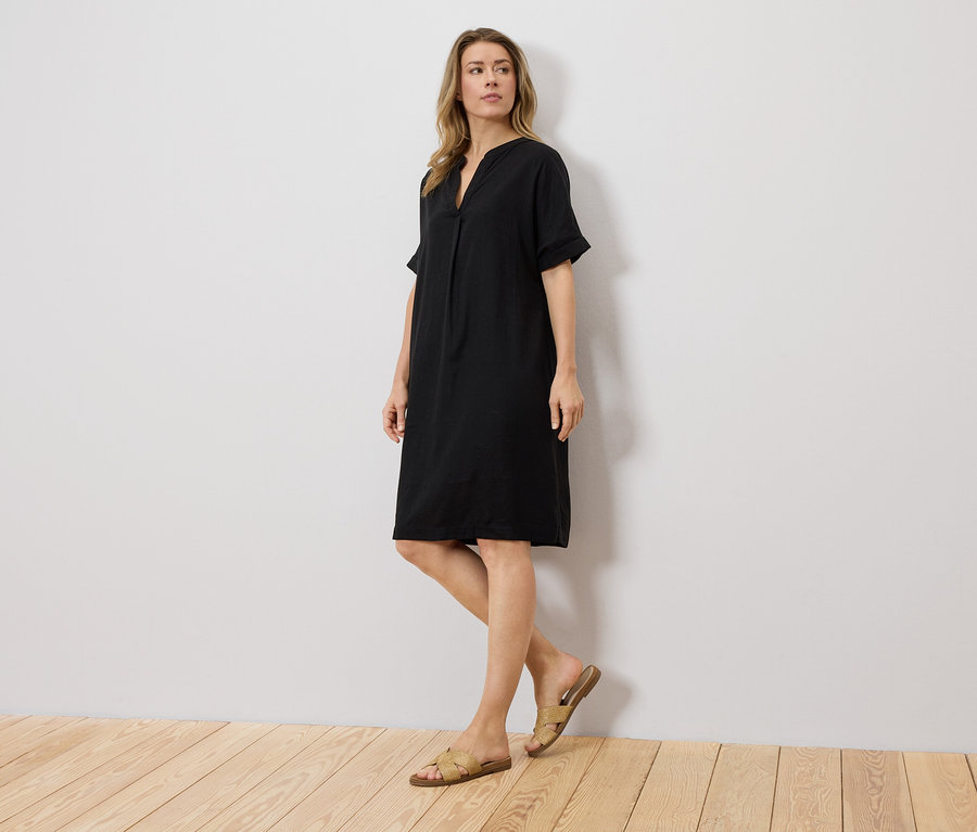 Frau steht vor einer Wand und trägt ein schwarzes Kleid und Sandalen.