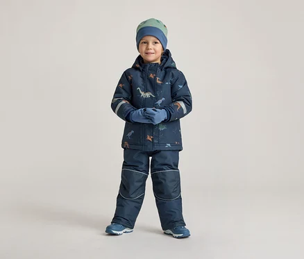 Ein Junge in marineblauer Winterkleidung mit Dinosaurier-Muster, Mütze und Handschuhen steht vor einem hellen Hintergrund.