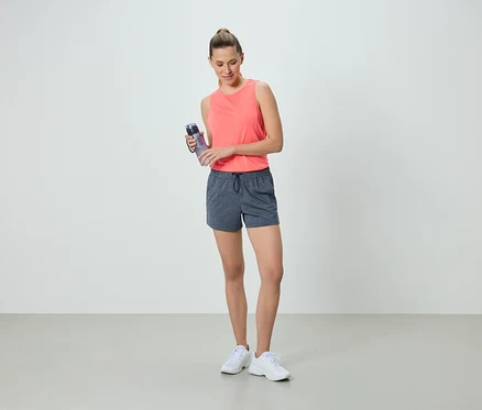 Frau in korallfarbenem Tanktop und grauen Shorts hält eine Wasserflasche.