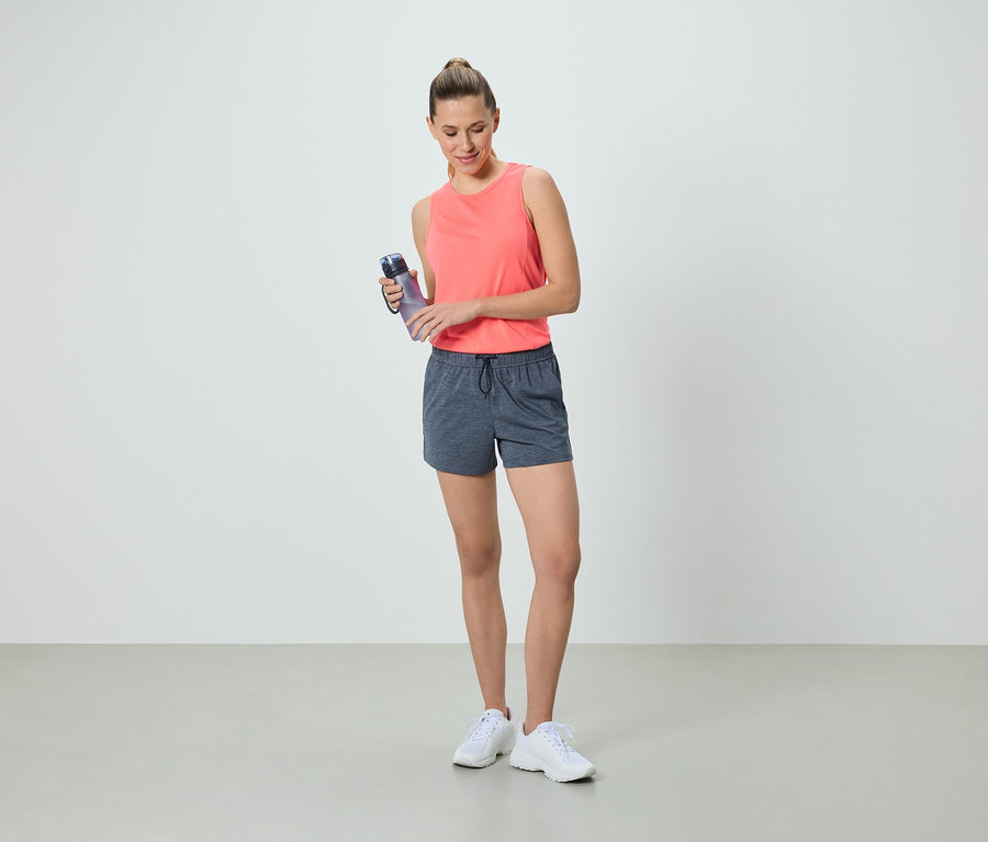Frau in korallfarbenem Tanktop und grauen Shorts hält eine Wasserflasche.