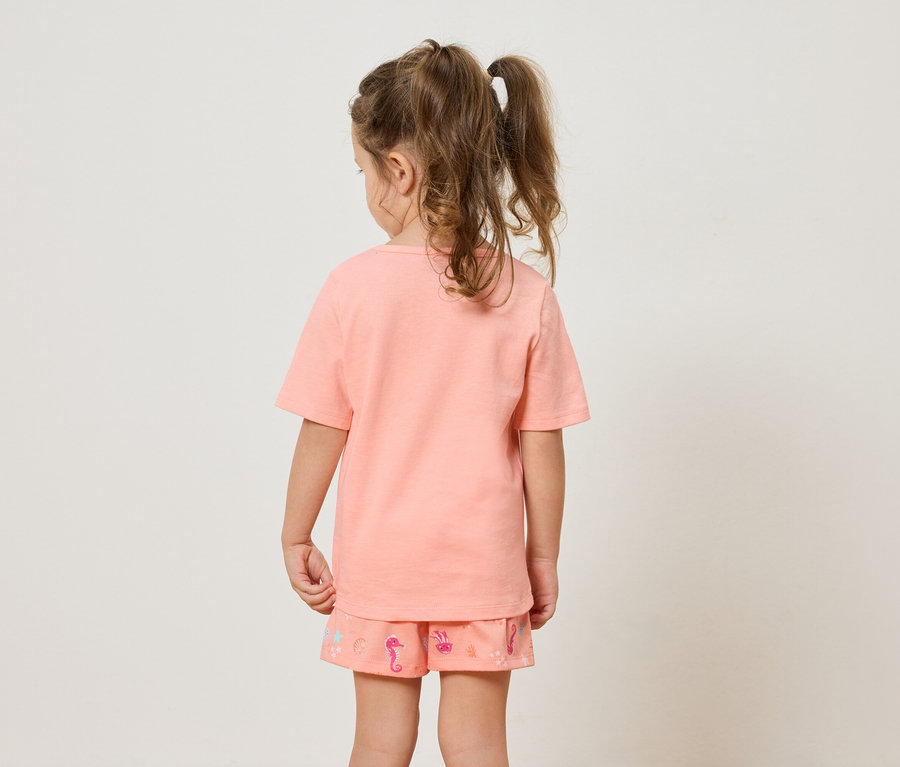 Rückansicht eines kleinen Mädchens mit Zöpfen, das ein orangefarbenes T-Shirt und Shorts mit Seepferdchen-Motiv trägt.