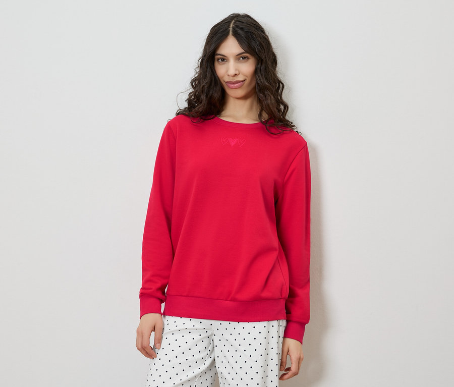 Eine Frau mit lockigem, braunem Haar trägt einen roten Pullover und eine gepunktete Hose vor einem weißen Hintergrund.