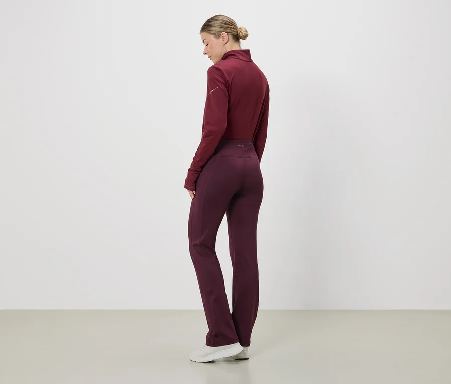 Eine Frau mit Dutt trägt ein weinrotes, langärmliges Oberteil und weinrote Leggings. Sie steht vor einer weißen Wand.
