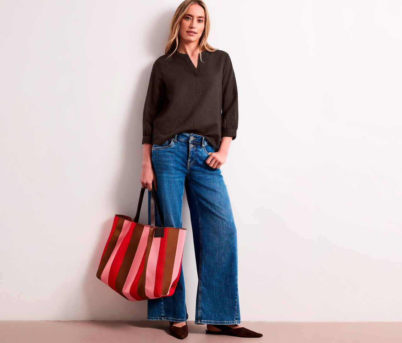 Eine Frau trägt eine dunkelgraue Street One Tunika-Bluse, weite Jeans und hält eine gestreifte Tragetasche in Rot, Pink und Braun.
