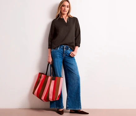 Eine Frau trägt eine dunkelgraue Street One Tunika-Bluse, weite Jeans und hält eine gestreifte Tragetasche in Rot, Pink und Braun.