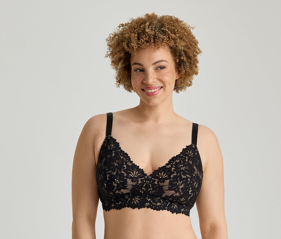 Eine lächelnde Frau mit hellbraunen Locken trägt einen schwarzen Spitzen-Bralette vor einem hellgrauen Hintergrund.