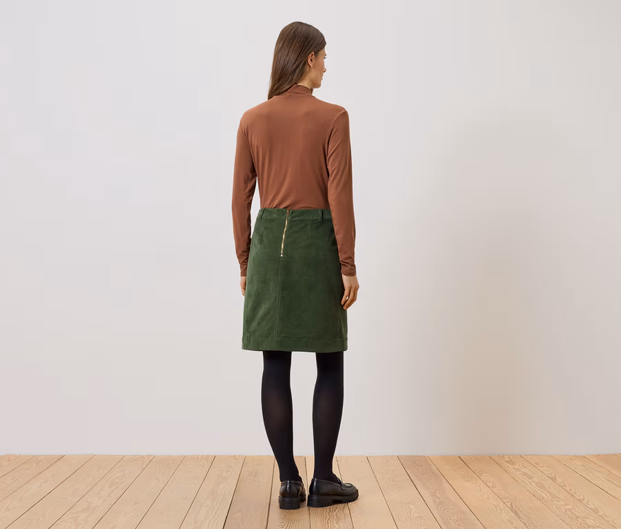 Eine Frau steht mit dem Rücken zur Kamera, trägt ein braunes Langarmshirt, einen grünen Rock, schwarze Strumpfhosen und schwarze Schuhe.