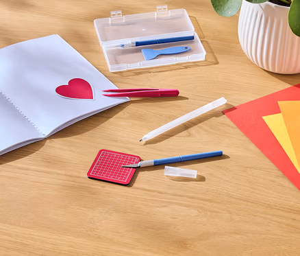 Auf einem Holztisch liegen ein offenes Notizbuch mit einem roten Herzaufkleber, eine Pinzette, ein Bastelmesser, eine Schneidematte und farbiges Papier.