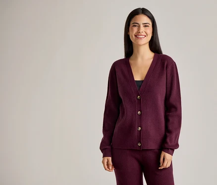 Lächelnde Frau mit langen, dunklen Haaren, die einen burgunderfarbenen Cardigan mit Knöpfen und eine Hose trägt.