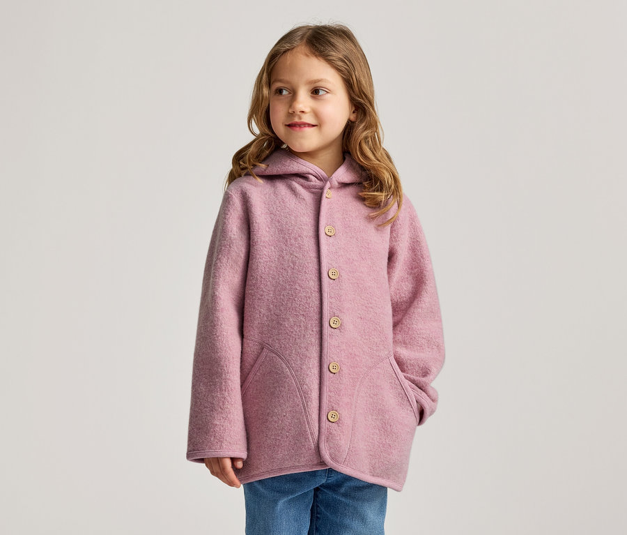 Ein junges Mädchen mit langen, gewellten braunen Haaren trägt eine rosa Kapuzenjacke mit Holzknöpfen und blaue Jeans. Sie blickt nach rechts.