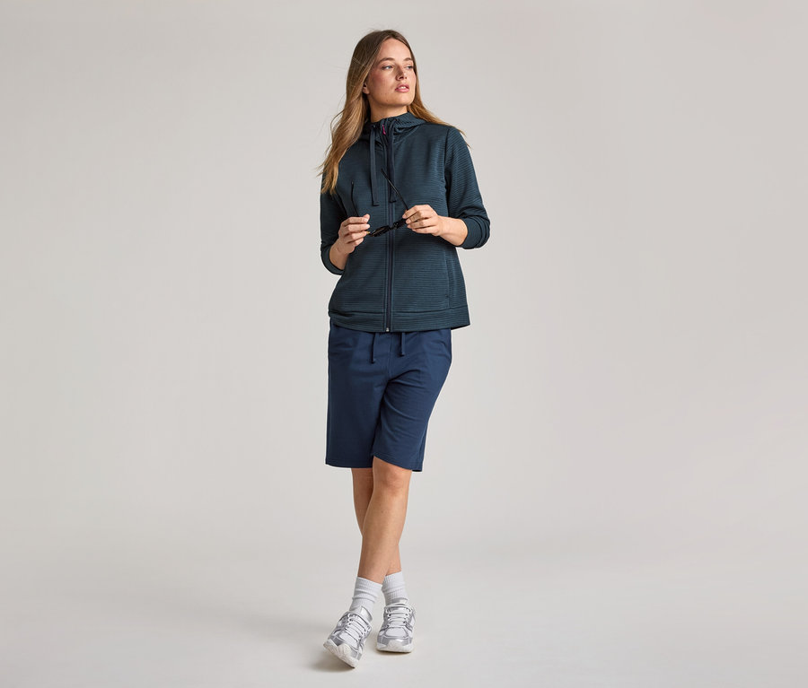 Frau mit Kapuzenjacke in Navy und Shorts. Sie hält eine Sonnenbrille in der Hand.