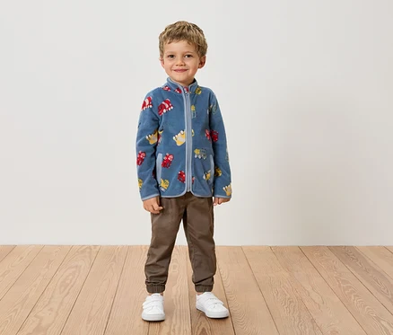 Ein Junge steht in einer blauen Fleecejacke mit Fahrzeugmuster, braunen Hosen und weißen Schuhen vor einer Wand.
