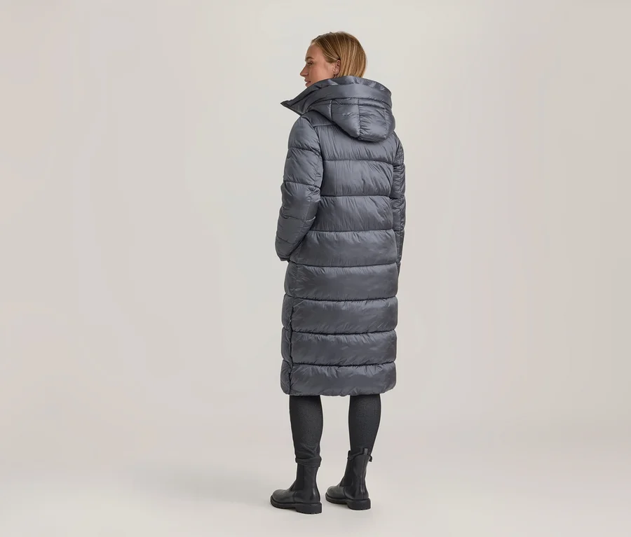 Frau von hinten, leicht gedreht, trägt einen langen, grauen Steppmantel mit Kapuze, dunkle Hose und schwarze Stiefel.
