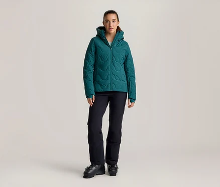 Frau steht in einer türkisfarbenen Steppjacke mit Kapuze, schwarzen Skihosen und schwarzen Skischuhen.