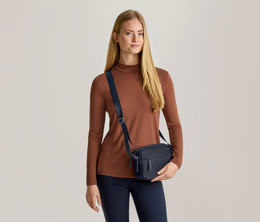 Eine Frau mit langen blonden Haaren trägt ein braunes Langarmshirt, eine dunkelblaue Hose und eine dunkelblaue Umhängetasche. Sie hält die Tasche mit ihrer linken Hand und blickt nach vorne.