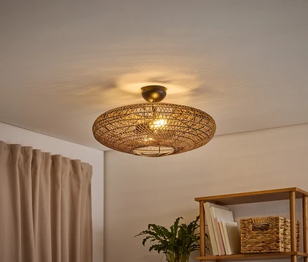 Eine ovale, geflochtene Deckenleuchte beleuchtet einen Raum und wirft Schatten an die Decke. Links hängt ein hellbrauner Vorhang, rechts steht ein Holzregal mit Büchern und einem geflochtenen Korb.