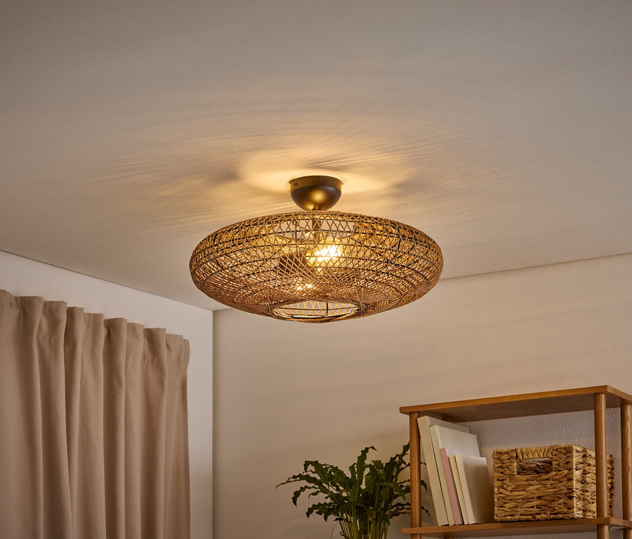 Eine ovale, geflochtene Deckenleuchte beleuchtet einen Raum und wirft Schatten an die Decke. Links hängt ein hellbrauner Vorhang, rechts steht ein Holzregal mit Büchern und einem geflochtenen Korb.