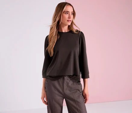 Eine Frau mit langen blonden Haaren trägt ein braunes Street One 3/4-Jerseyshirt und eine gemusterte Hose vor rosa Hintergrund.