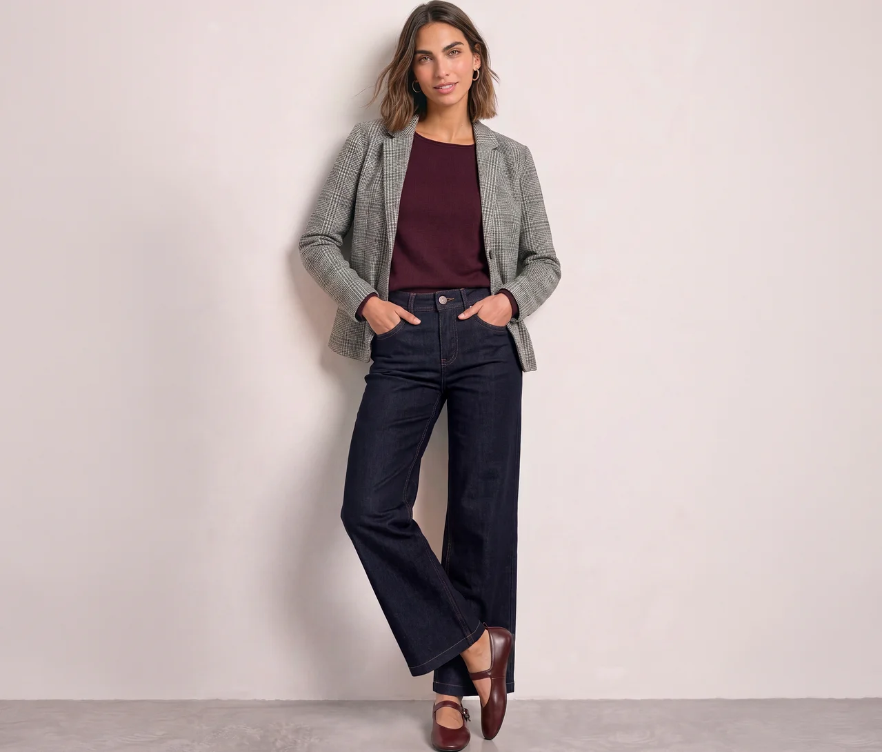 Eine Frau steht mit den Händen in den Taschen, trägt einen grau karierten Street One Blazer über einem bordeauxroten Oberteil, dunkelblaue weite Jeans und dunkelrote flache Schuhe, an eine helle Wand gelehnt.