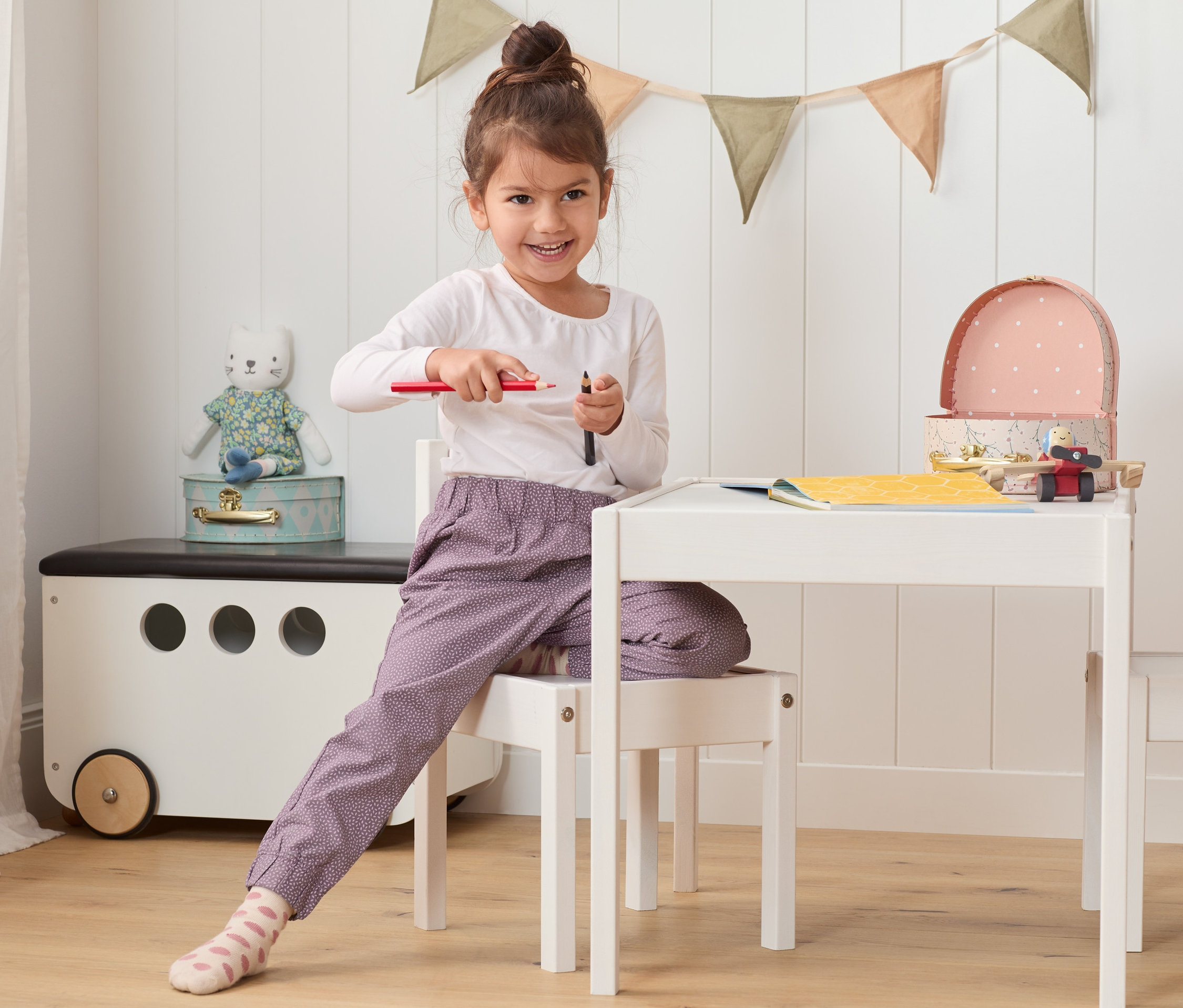 Mädchen sitzt am Tisch und hält Stifte. Sie trägt eine Baumwoll-Schlupfhose.