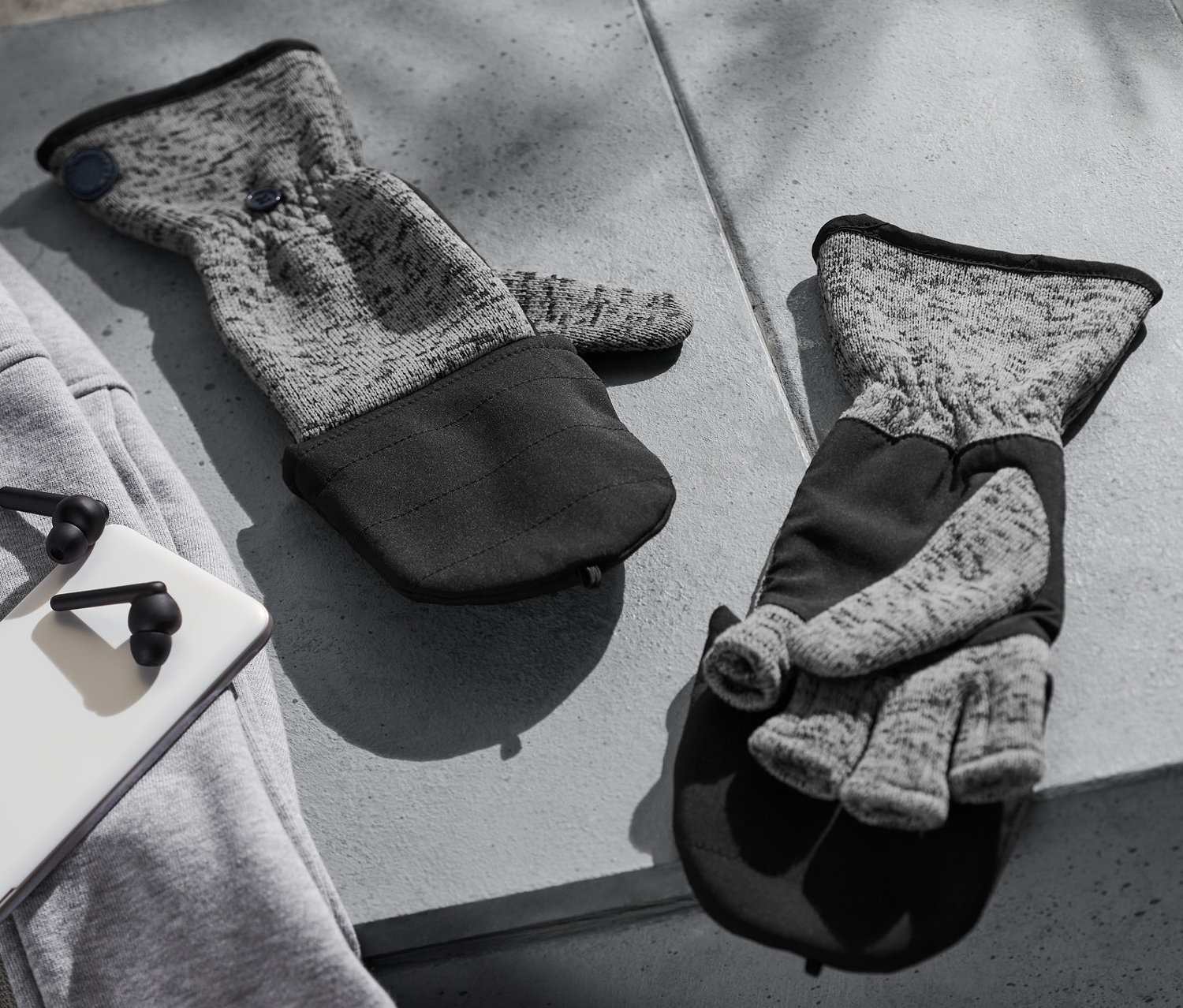 Zwei Strickfleece-Klappfäustlinge liegen auf einem grauen Betontisch neben einem grauen Sweatshirt, einem weißen Telefon und schwarzen Kopfhörern.