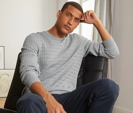 Mann sitzt auf einem schwarzen Ledersessel und trägt einen grau melierten Strickpullover.