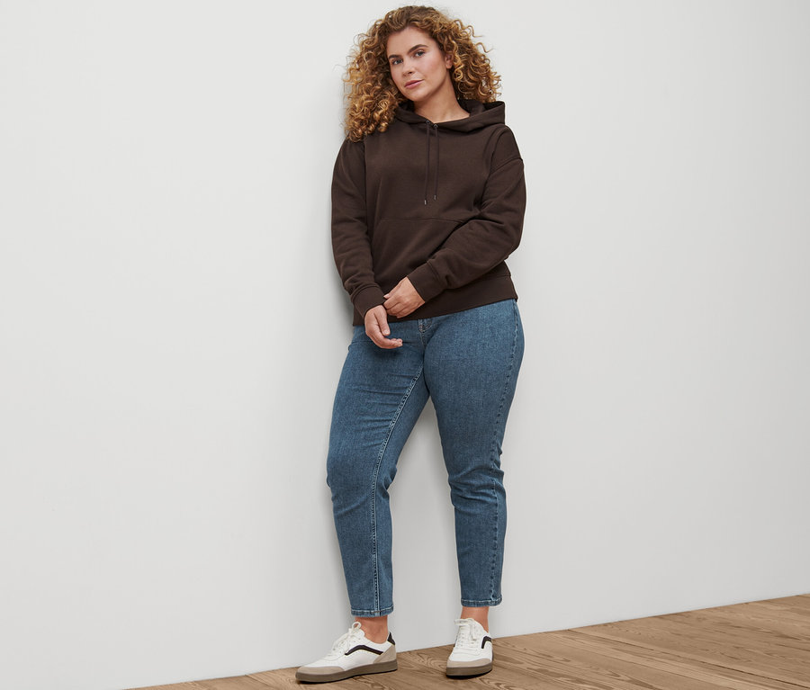 Eine Frau steht und trägt Straight-Denim »Fit Juna« in Midblue, Leder-Sneaker und einen dunkelbraunen Hoodie.