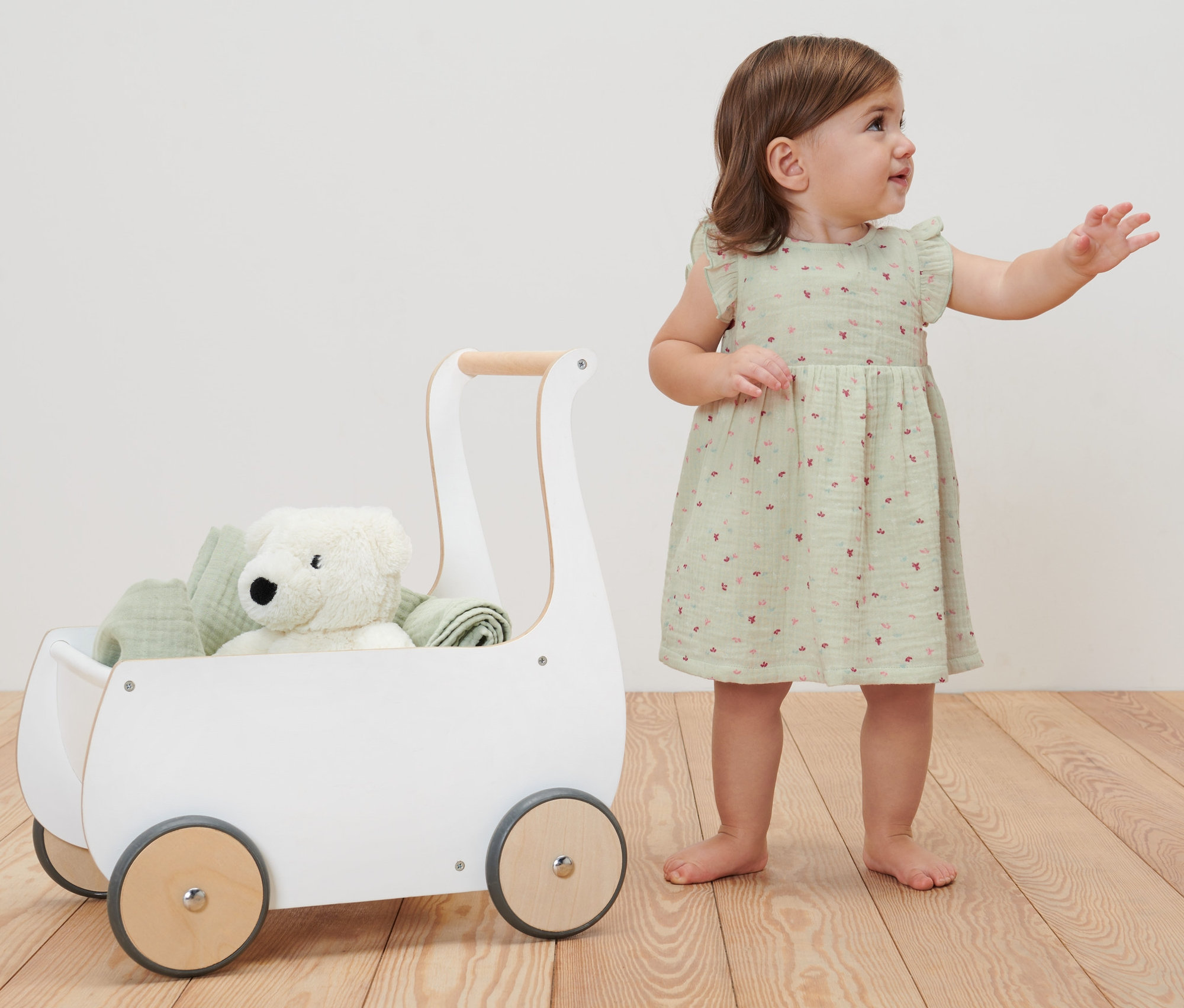 Kleinkind steht neben einem weißen Spielzeugwagen und trägt ein Baby-Webkleid.