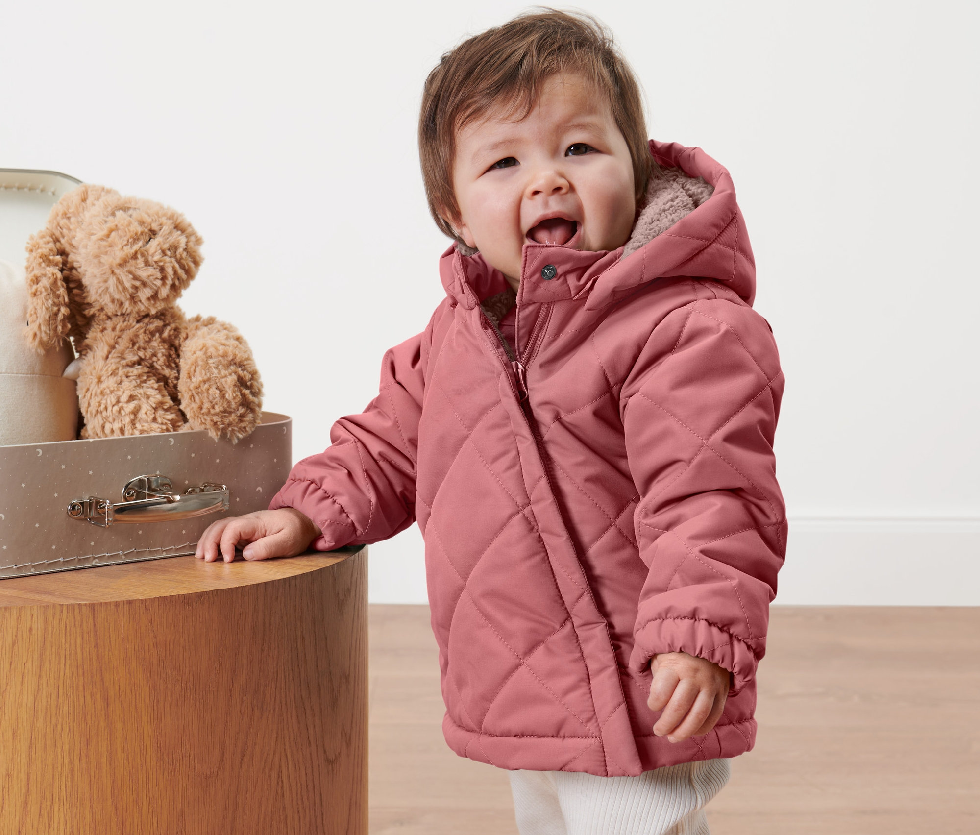 Ein Kind in einer rosa Kinder-Steppjacke steht neben einem Koffer mit Spielzeug.