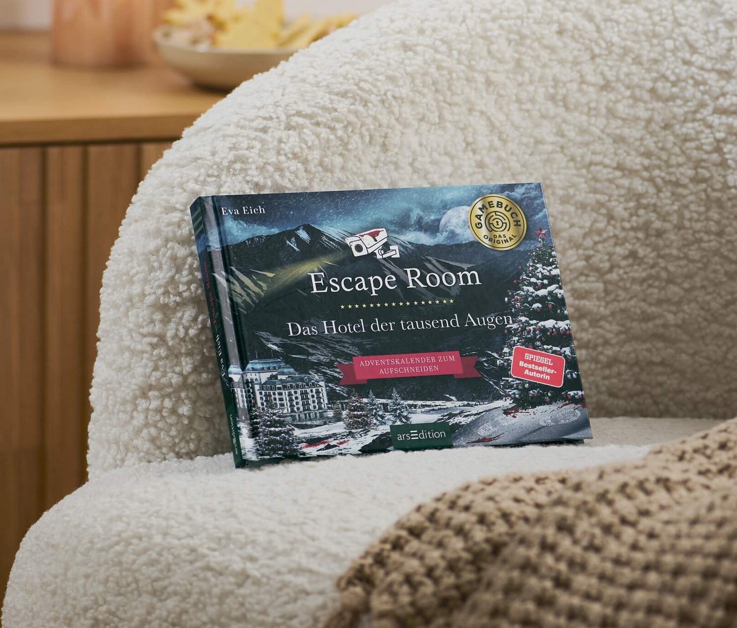 Auf einem Sessel liegen ein Escape-Room-Adventskalenderbuch »Das Hotel der tausend Augen« und eine beige Decke.