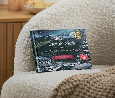 Auf einem Sessel liegen ein Escape-Room-Adventskalenderbuch »Das Hotel der tausend Augen« und eine beige Decke.
