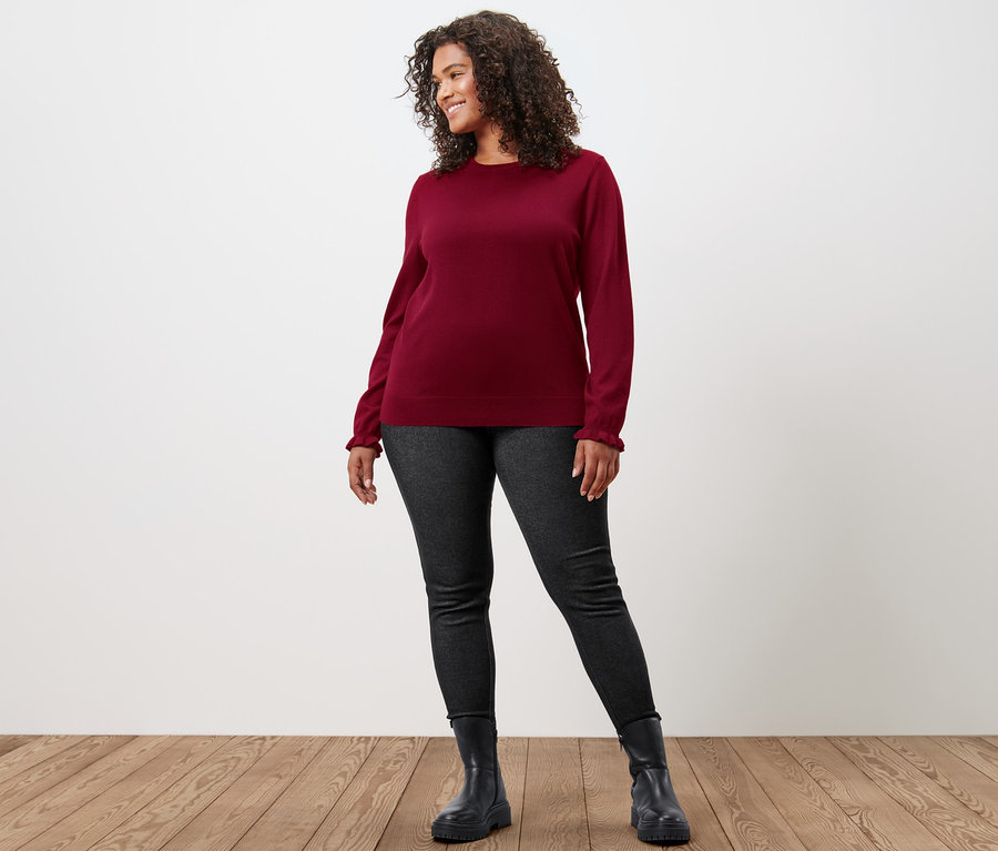 Frau posiert in bordeauxrotem Feinstrickpullover mit Rüschendetail und gefütterten Leggings in Jeansoptik.