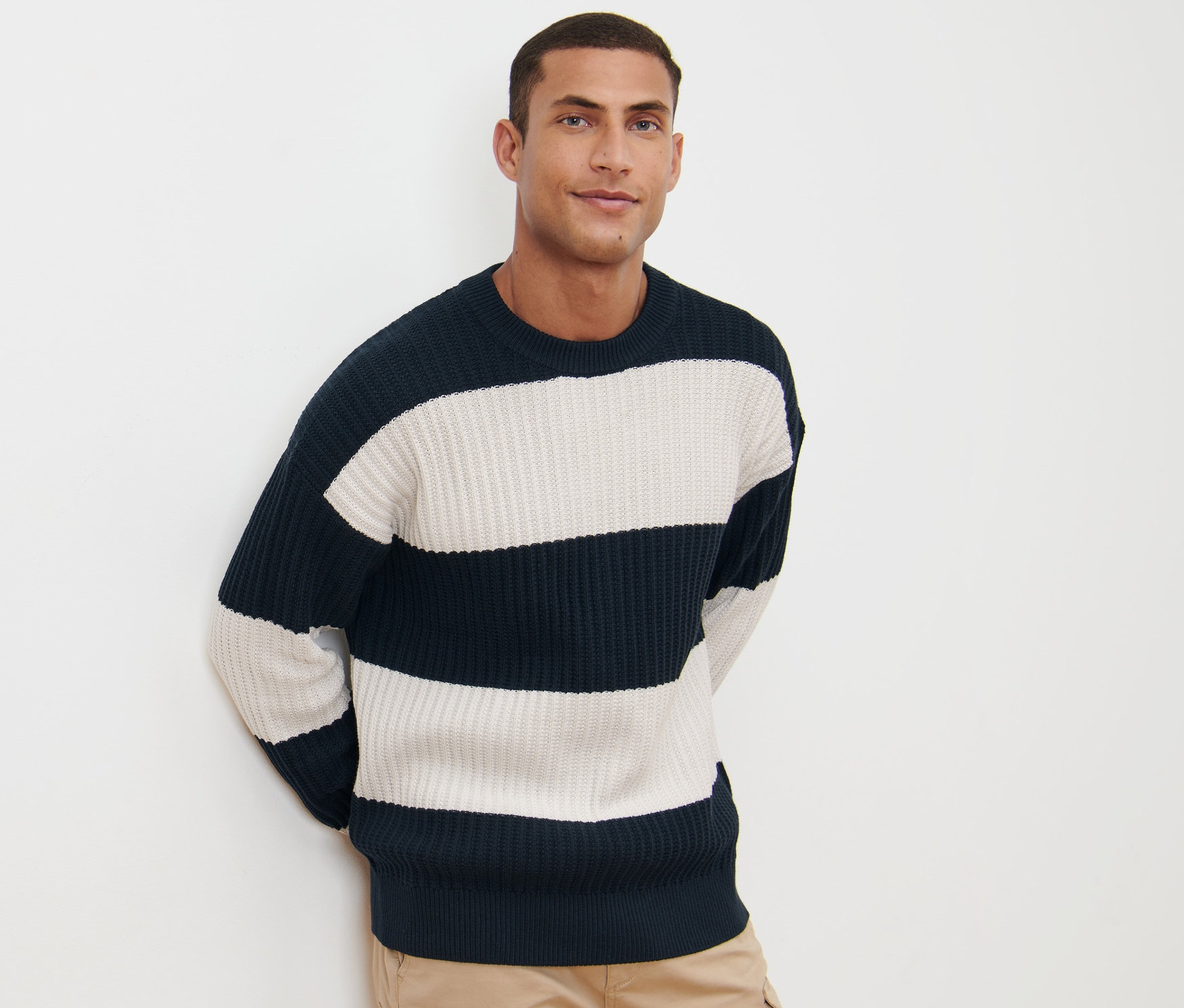 Mann posiert vor weißem Hintergrund mit einem gestreiften Grobstrick-Pullover in Marineblau und Weiß und Khaki-Hosen.