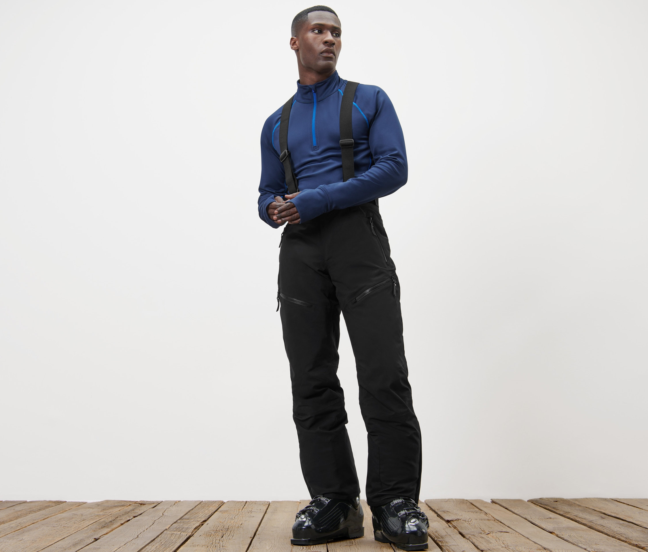 Mann steht auf Holzboden und trägt ein blaues Oberteil und eine High-Tech-Skihose.