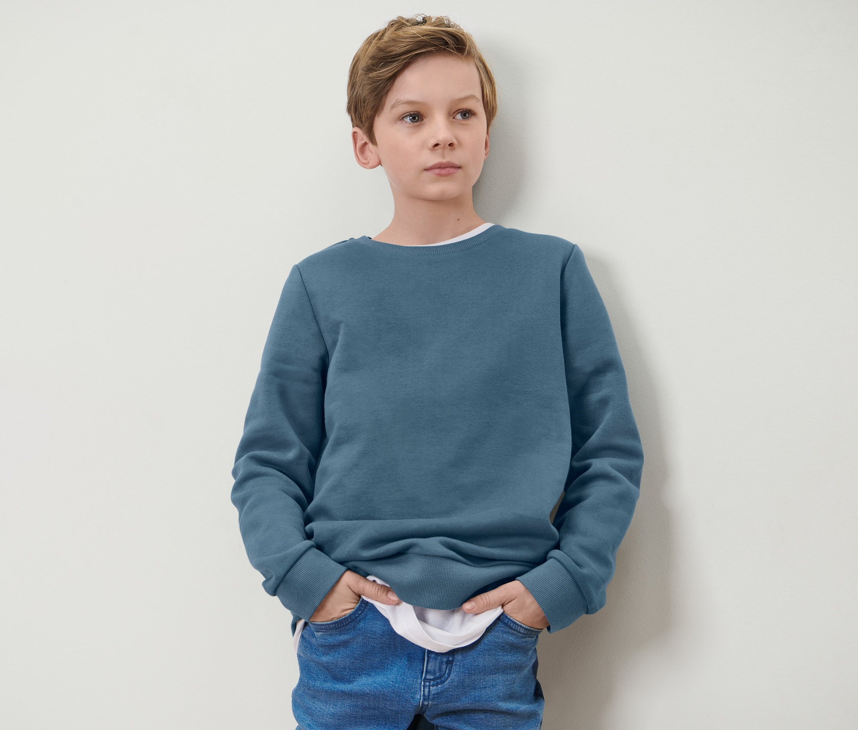 Junge steht mit Händen in den Taschen eines blauen Kinder-Sweatshirts.