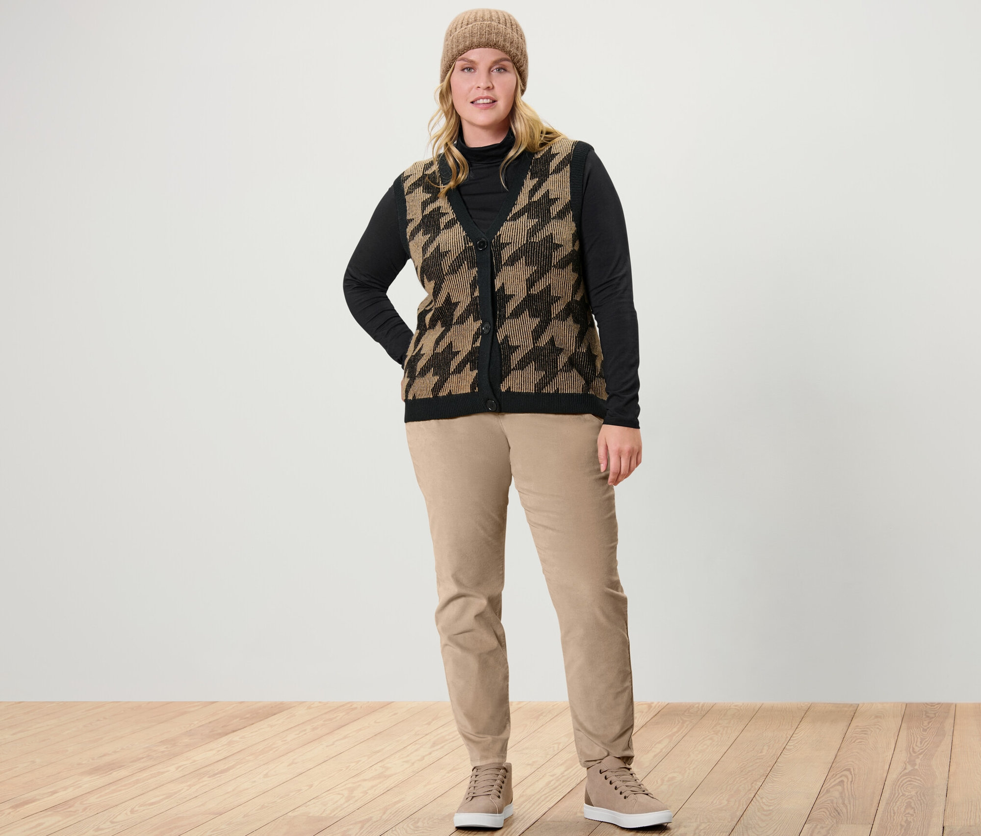 Eine Frau steht und trägt ein schwarzes Rollkragenshirt, eine Strickweste mit Hahnentritt-Jacquard und eine camel Samthose.