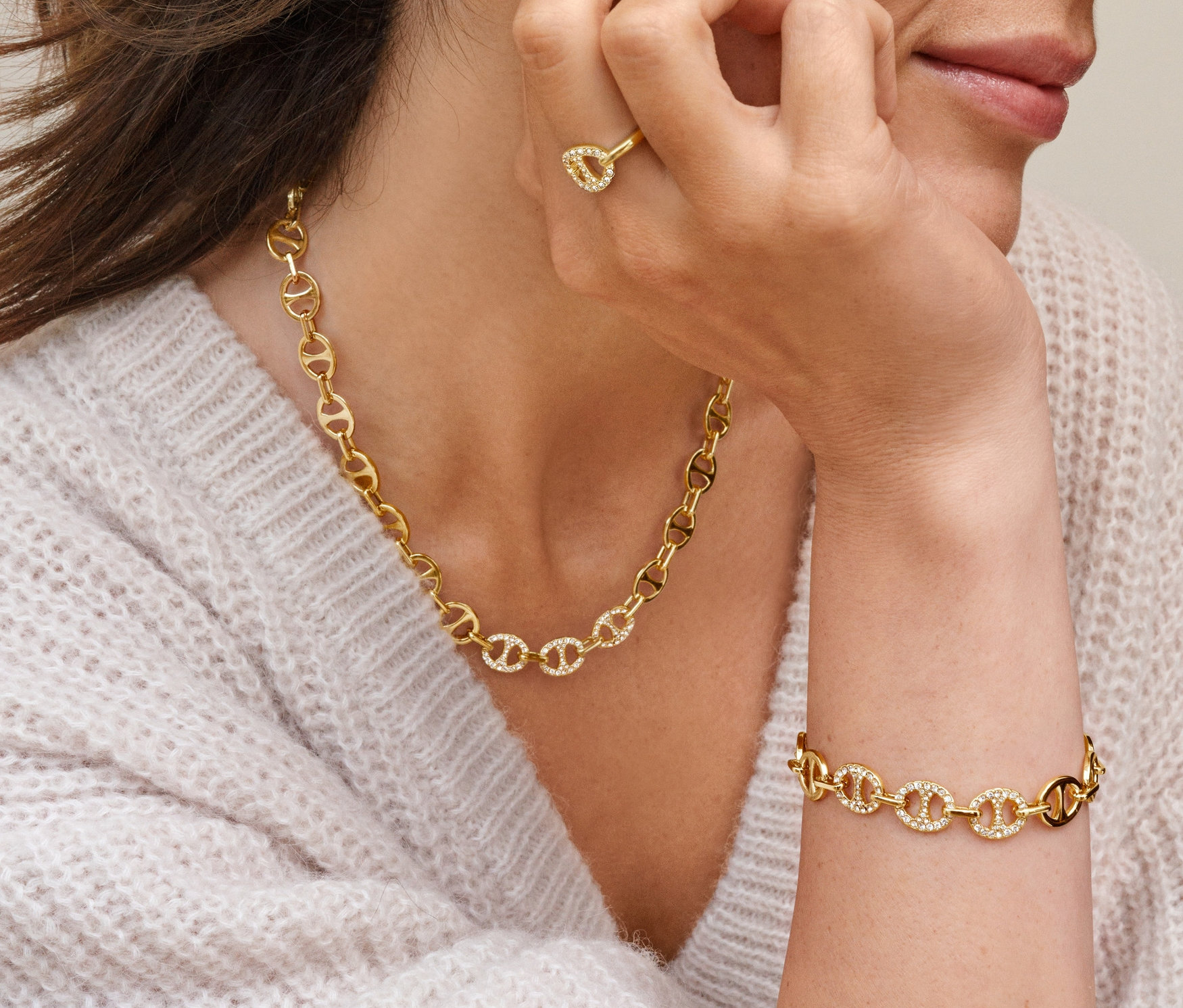 Frau mit geschlossenen Augen berührt ihr Ohr, trägt ovale Creolen mit Glaskristallen, Ring Schiffsanker mit Glaskristallen, Kette Schiffsanker mit Glaskristallen und Armband Schiffsanker mit Glaskristallen.