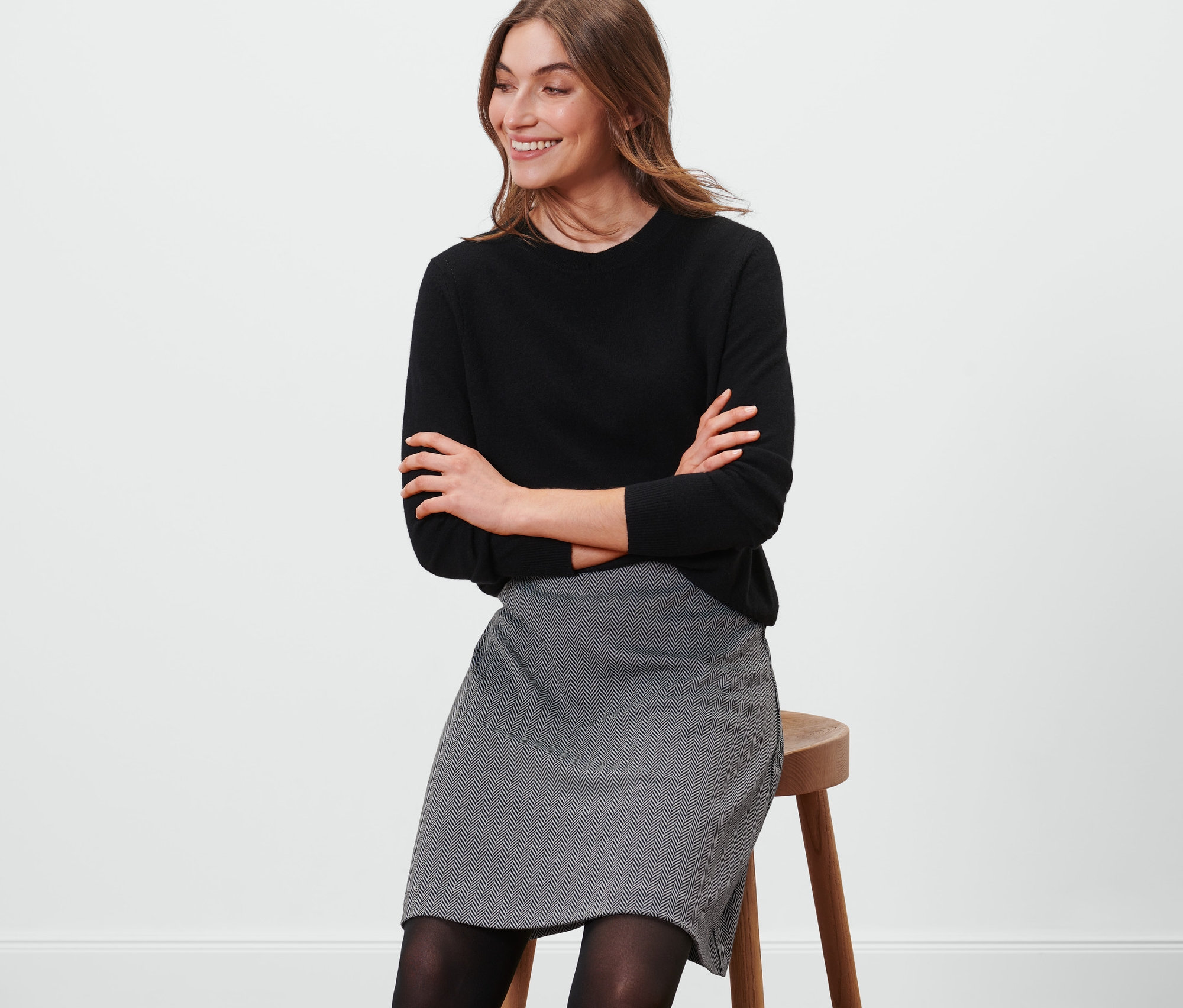 Frau sitzt auf einem Hocker und trägt einen schwarzen Pullover und einen Sweatrock.