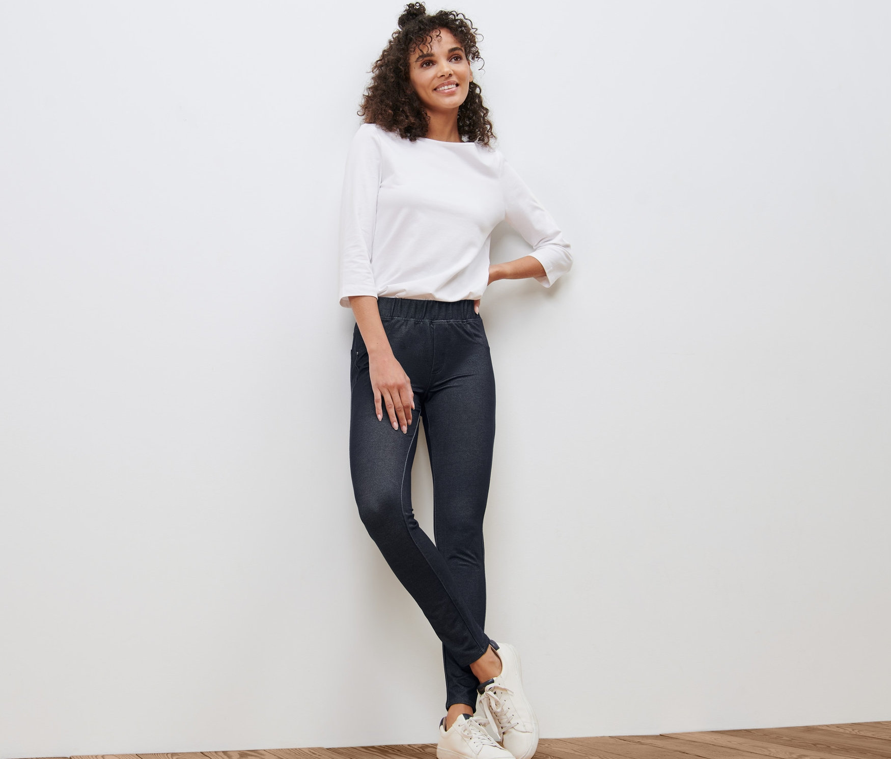 Frau, die an einer weißen Wand lehnt, trägt Leggings in Jeansoptik, eine weiße Bluse und Turnschuhe.