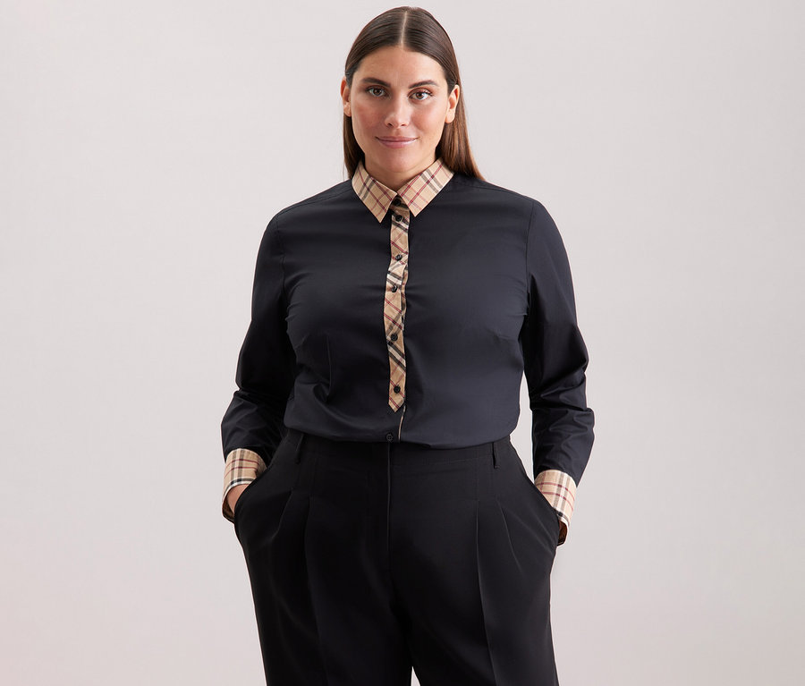 Frau mit braunen Haaren trägt eine schwarze Seidensticker Popeline-Bluse mit Karodetail und eine schwarze Hose.