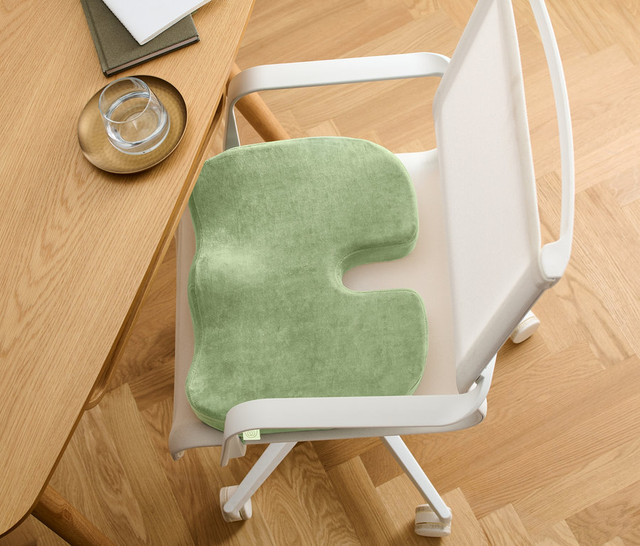 Grünes Sitzkissen mit U-förmigem Ausschnitt auf einem Stuhl. Der Stuhl steht an einem Holztisch.