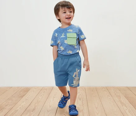 Junge steht und trägt blaue Kinder-Sweatshorts und ein T-Shirt.