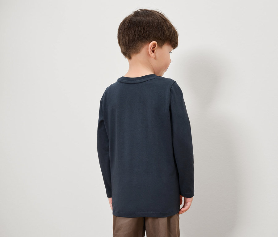 Ein Junge mit dem Rücken zur Kamera trägt ein dunkelblaues Langarmshirt und braune Hosen.
